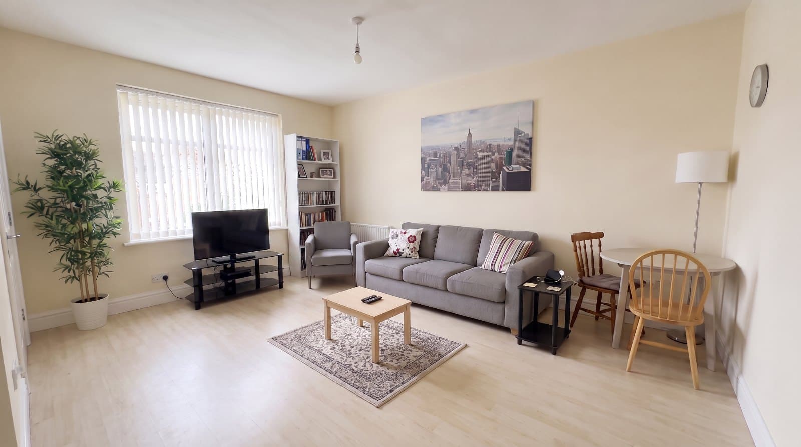 An ordinary living room photographed by its owner — a grey flat-pack sofa, a too-small rug, vertical blinds and one bare ceiling bulb